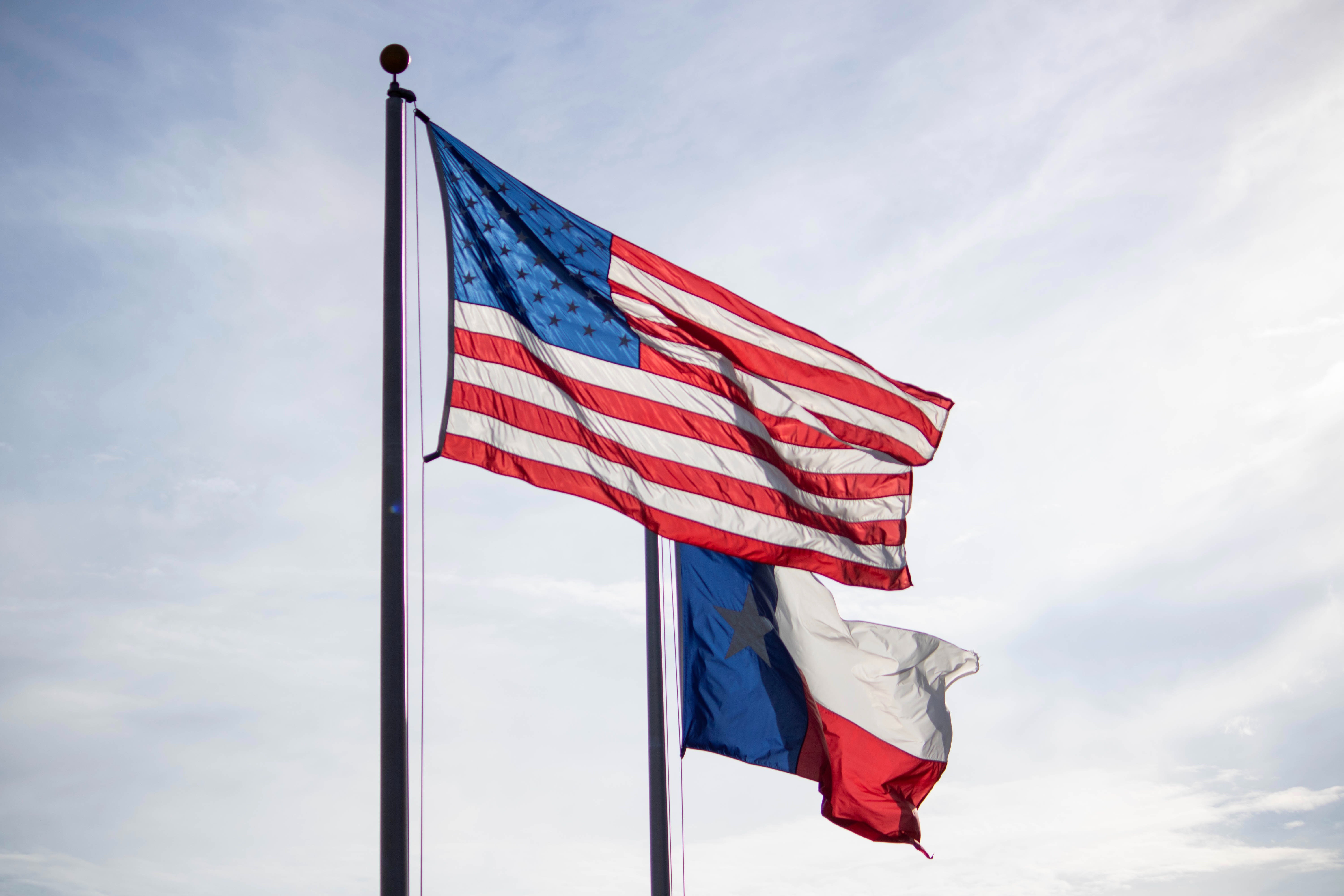 United States and Texas flags flying with a sunny sky in the background.